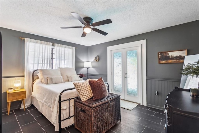 bedroom featuring a ceiling fan, french doors, a textured ceiling, a wainscoted wall, and access to outside
