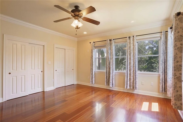 unfurnished bedroom featuring crown molding, two closets, a ceiling fan, baseboards, and wood finished floors