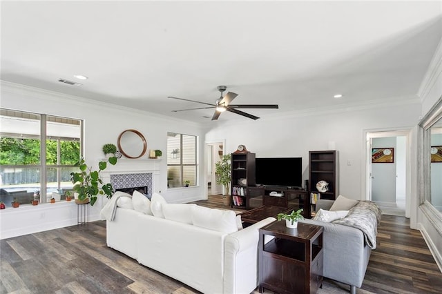 living room with crown molding, a tiled fireplace, a ceiling fan, and dark wood finished floors