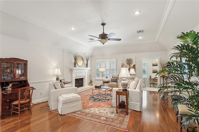 living room featuring a ceiling fan, visible vents, wood finished floors, a fireplace with raised hearth, and ornamental molding