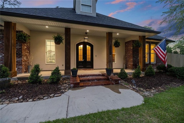 exterior entry at dusk featuring a porch, french doors, roof with shingles, brick siding, and stucco siding