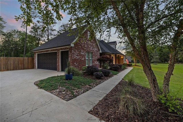 property exterior at dusk with driveway, brick siding, fence, a yard, and a shingled roof
