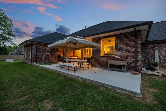 back of house at dusk featuring brick siding, a patio area, roof with shingles, and a yard