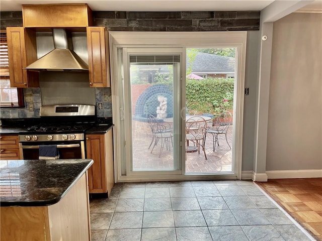 kitchen featuring backsplash, brown cabinets, stainless steel gas stove, baseboards, and wall chimney exhaust hood
