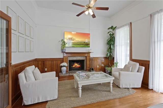 living area with wood walls, wainscoting, wood finished floors, ceiling fan, and a lit fireplace