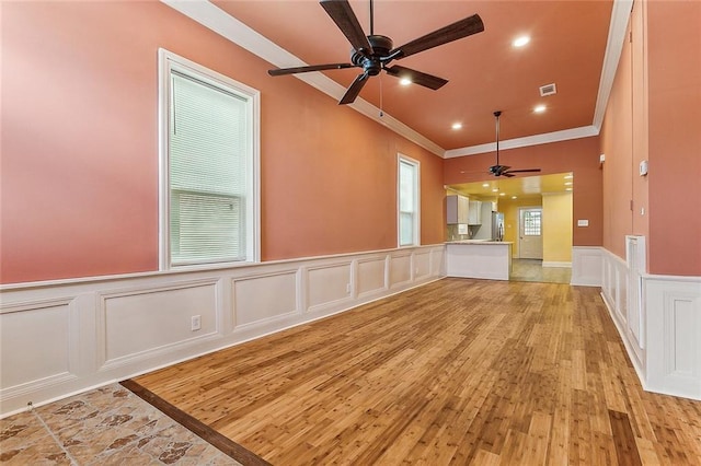 unfurnished living room featuring ceiling fan, visible vents, wainscoting, ornamental molding, and a decorative wall