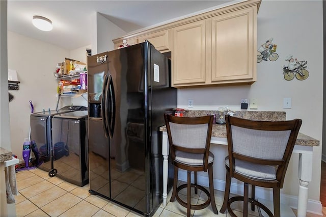 kitchen with black fridge with ice dispenser, a kitchen bar, cream cabinetry, washing machine and clothes dryer, and light tile patterned floors