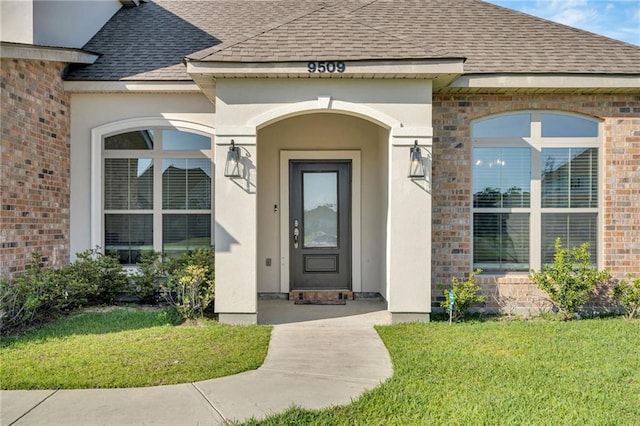 property entrance featuring a lawn, brick siding, roof with shingles, and stucco siding