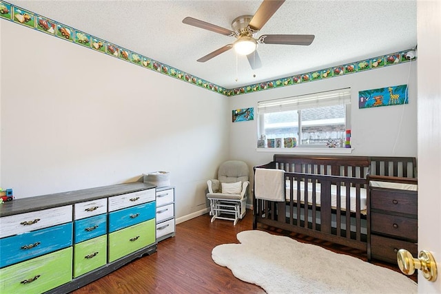 bedroom with wood finished floors, baseboards, a textured ceiling, a crib, and a ceiling fan