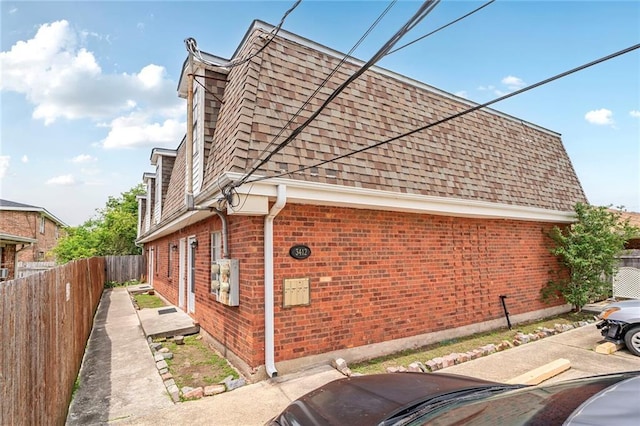 view of side of home featuring mansard roof, brick siding, a shingled roof, and fence