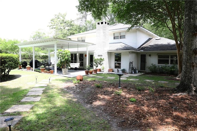 rear view of property featuring a chimney, a ceiling fan, a lawn, brick siding, and a patio