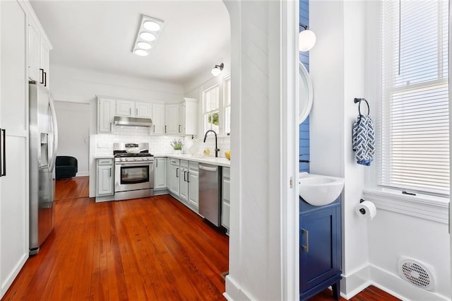 kitchen featuring stainless steel appliances, under cabinet range hood, dark wood-style flooring, light countertops, and tasteful backsplash
