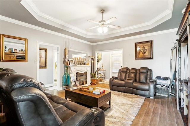 living room featuring a raised ceiling, wood finished floors, a ceiling fan, a fireplace, and crown molding