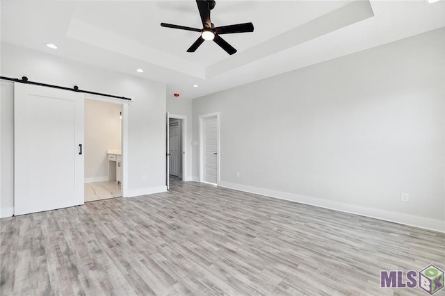 unfurnished bedroom featuring light wood finished floors, a raised ceiling, a barn door, and recessed lighting
