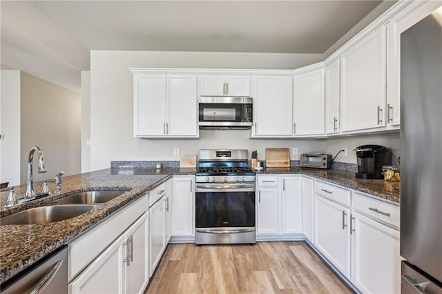 kitchen featuring appliances with stainless steel finishes, a sink, white cabinets, dark stone counters, and light wood-style floors