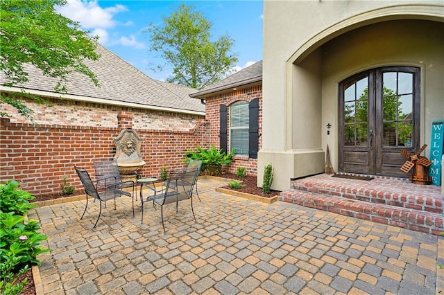 property entrance featuring brick siding, french doors, roof with shingles, a patio area, and stucco siding
