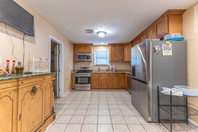kitchen featuring stainless steel appliances, brown cabinets, light tile patterned floors, a sink, and dark stone countertops