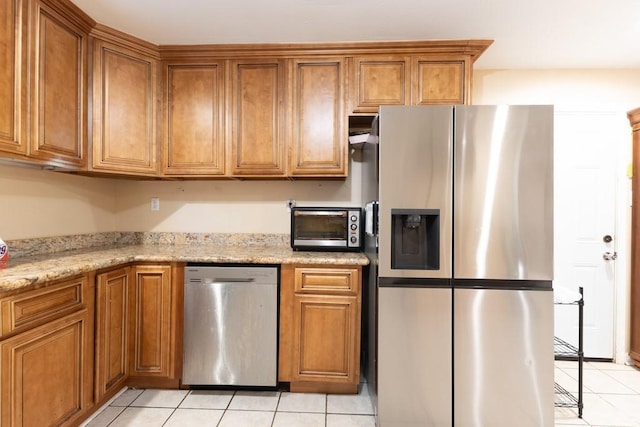 kitchen featuring appliances with stainless steel finishes, light tile patterned floors, brown cabinetry, and light stone counters