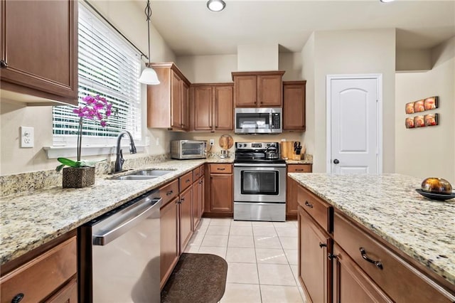 kitchen featuring stainless steel appliances, a sink, brown cabinets, decorative light fixtures, and light stone counters