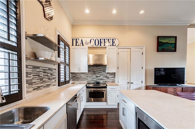 kitchen with appliances with stainless steel finishes, ornamental molding, wall chimney exhaust hood, backsplash, and white cabinetry