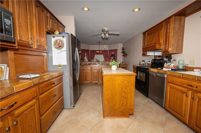 kitchen with stainless steel appliances, a center island, a ceiling fan, brown cabinetry, and tile counters