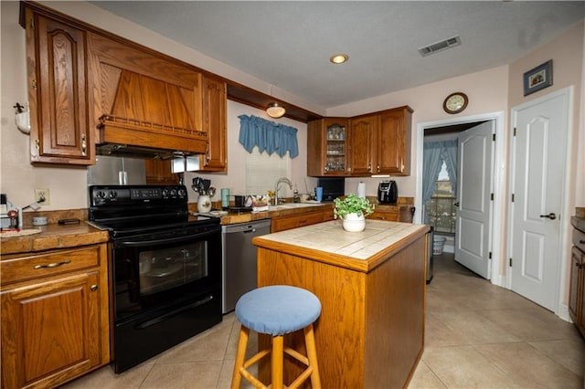 kitchen featuring black electric range oven, dishwasher, tile counters, brown cabinetry, and custom range hood