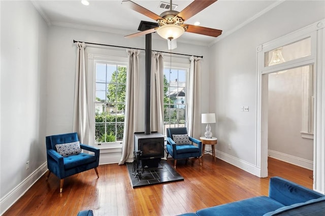 living area with a wood stove, wood-type flooring, baseboards, ornamental molding, and a ceiling fan