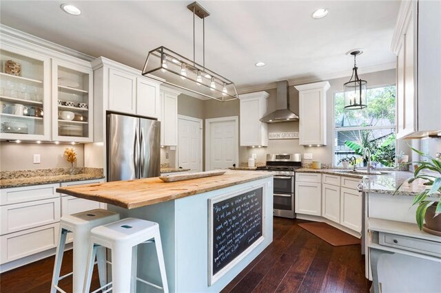 kitchen featuring stainless steel appliances, wall chimney range hood, a sink, butcher block countertops, and white cabinets
