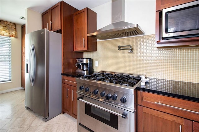 kitchen featuring appliances with stainless steel finishes, wall chimney range hood, backsplash, dark stone counters, and light tile patterned floors