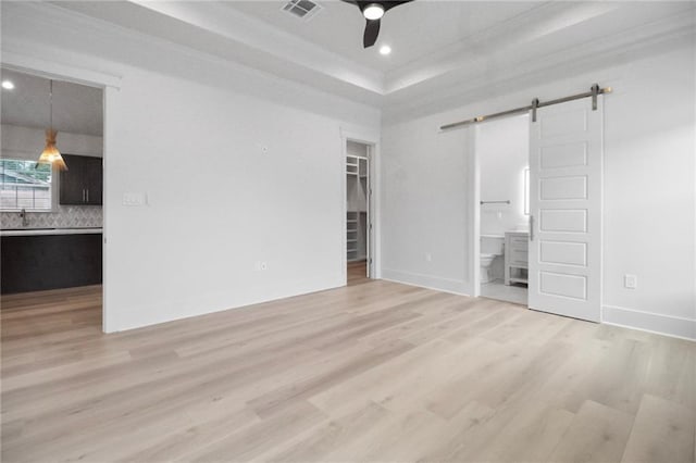 unfurnished bedroom with a barn door, a walk in closet, connected bathroom, light wood finished floors, and a tray ceiling