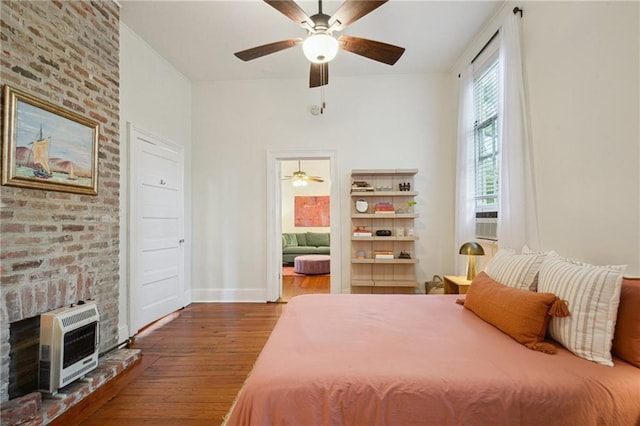 bedroom featuring heating unit, wood finished floors, a fireplace, baseboards, and a ceiling fan