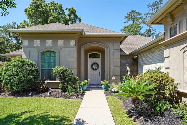 view of exterior entry featuring a shingled roof, a lawn, and stucco siding