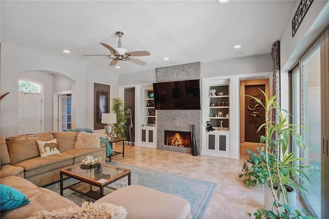 living room featuring a ceiling fan, arched walkways, recessed lighting, a tiled fireplace, and built in features