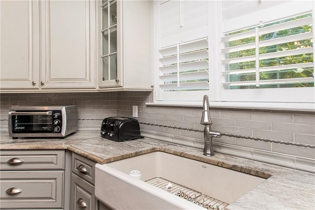 kitchen with a sink, tasteful backsplash, glass insert cabinets, and light stone counters