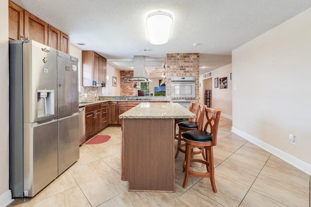 122 Warwick St, La Place, LA 70068 - kitchen featuring appliances with stainless steel finishes, island range hood, brown cabinetry, light tile patterned floors, and light stone countertops