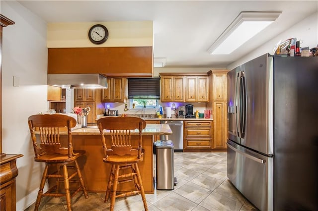 1500 Madewood Rd, La Place, LA 70068 - kitchen featuring stainless steel appliances, a peninsula, wall chimney exhaust hood, a kitchen bar, and brown cabinets