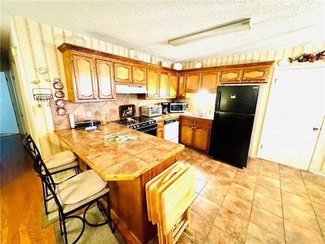 6655 Dalark Dr, Baton Rouge, LA 70812 - kitchen featuring black appliances, under cabinet range hood, a peninsula, brown cabinets, and a textured ceiling