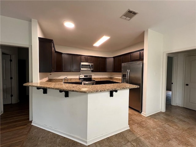 500 Buckthorne Pl, Covington, LA 70435 - kitchen featuring appliances with stainless steel finishes, a peninsula, dark brown cabinetry, light stone countertops, and a kitchen bar