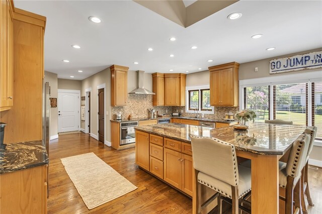 kitchen with wall chimney range hood, stainless steel appliances, decorative backsplash, and wood finished floors