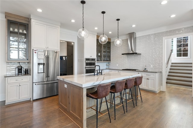 kitchen with decorative backsplash, stainless steel appliances, wall chimney range hood, a center island with sink, and white cabinets