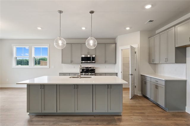 kitchen featuring pendant lighting, gray cabinetry, stainless steel appliances, wood-type flooring, and a center island with sink