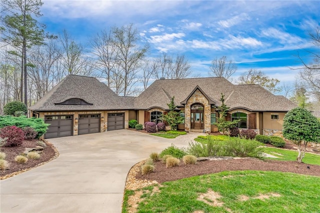 french country style house featuring an attached garage, stone siding, driveway, roof with shingles, and stucco siding