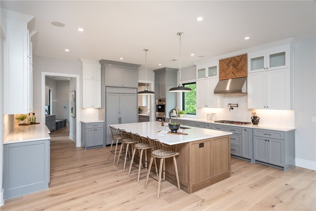 kitchen with light wood-type flooring, gray cabinetry, a large island with sink, and wall chimney range hood