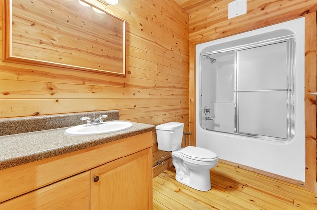 full bathroom featuring wood-type flooring, shower / bath combination with glass door, toilet, wooden walls, and vanity