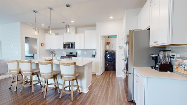 kitchen featuring appliances with stainless steel finishes, white cabinetry, an island with sink, wood-type flooring, and washer / dryer