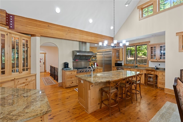 kitchen featuring pendant lighting, high vaulted ceiling, light hardwood / wood-style flooring, a center island with sink, and light stone countertops