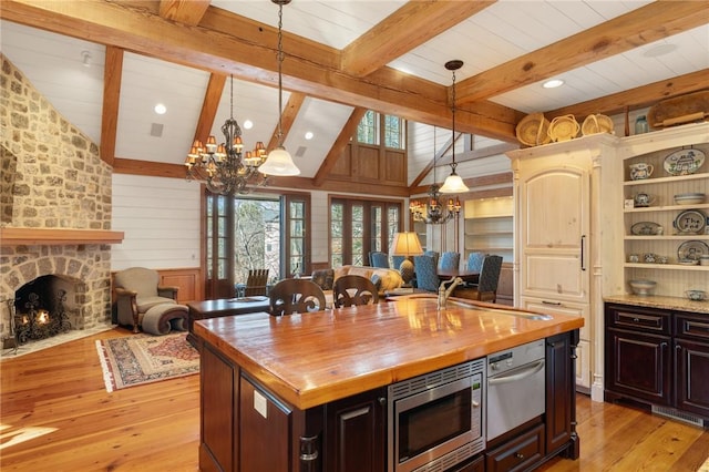 kitchen with a stone fireplace, a kitchen island with sink, a chandelier, stainless steel microwave, and light hardwood / wood-style flooring