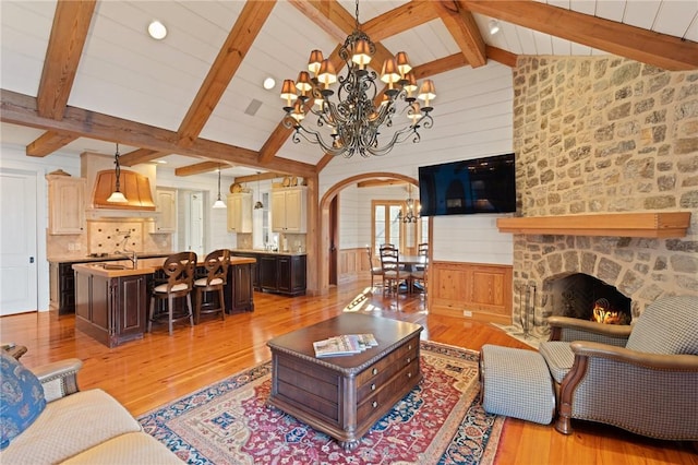 living room with beamed ceiling, an inviting chandelier, a stone fireplace, and light wood-type flooring