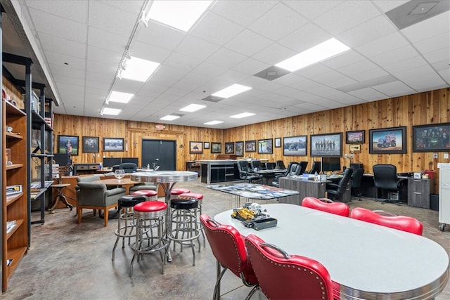 dining room with a drop ceiling, wooden walls, and concrete floors