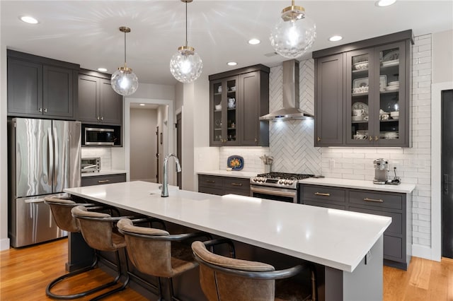 kitchen featuring hanging light fixtures, appliances with stainless steel finishes, a kitchen island with sink, and wall chimney exhaust hood